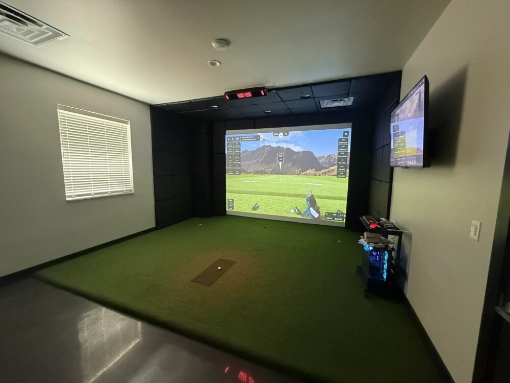Bright home golf simulator room with black enclosure, turf floor and wall-mounted display